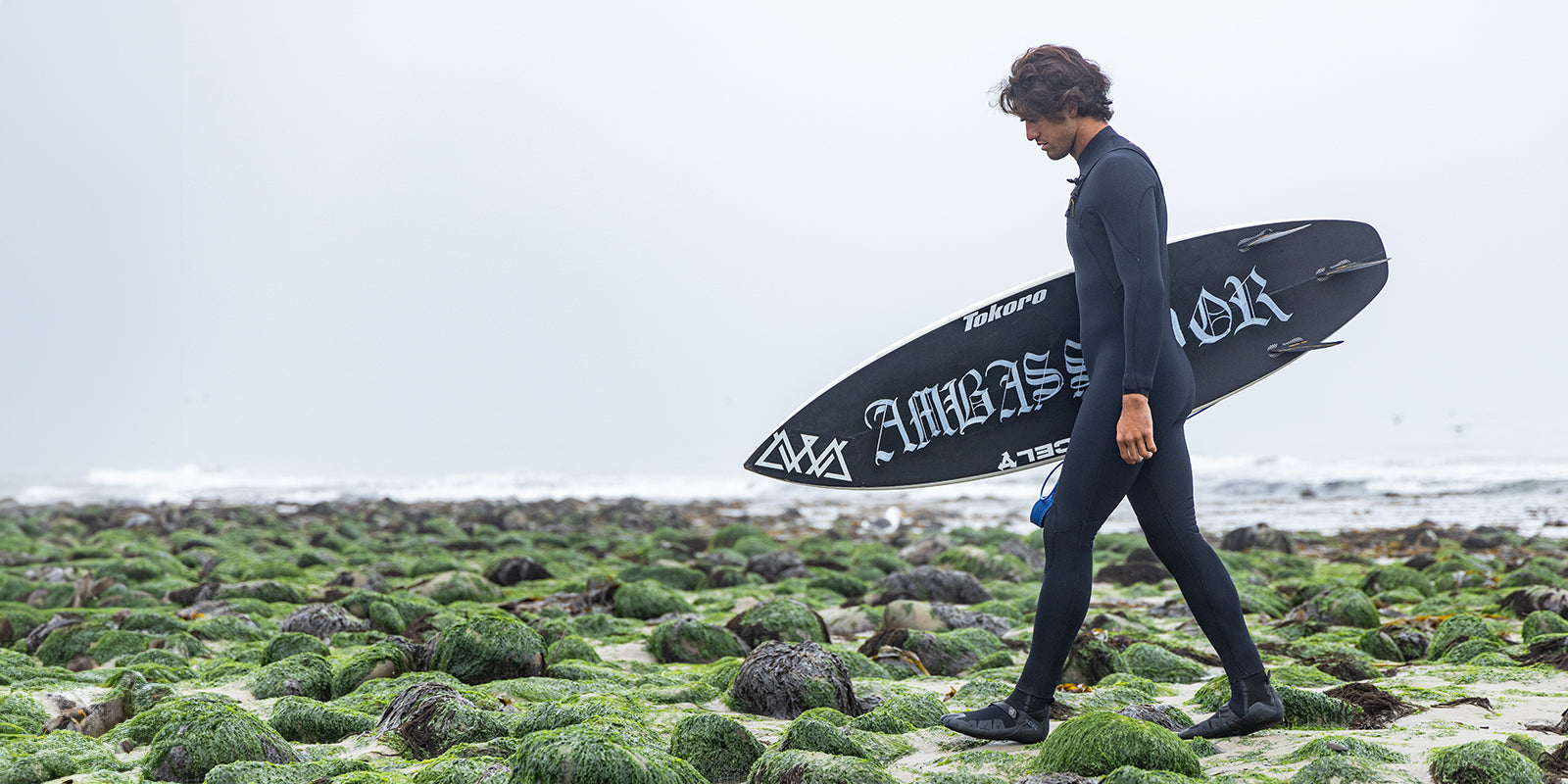 Zeke Lau walking in his Xcel wetsuit on a rocky beach holding his surfboard 